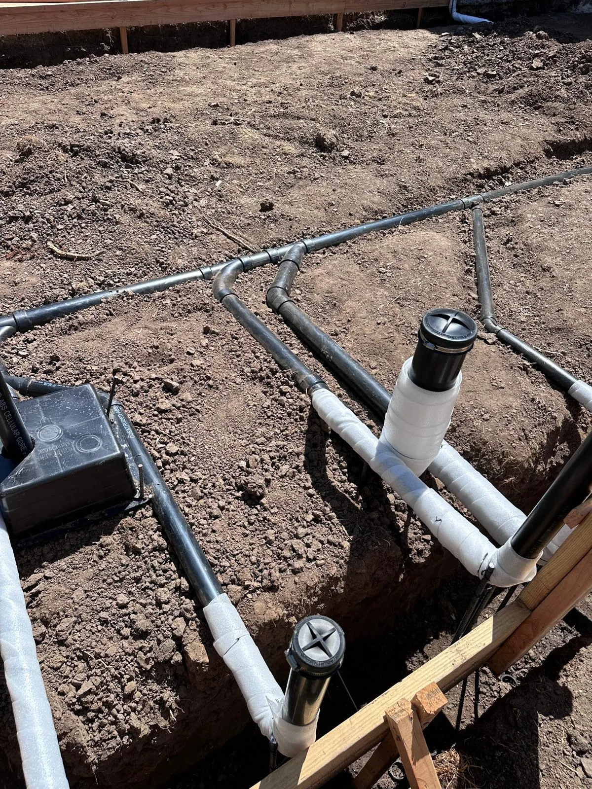 Black drain lines with wrapped fittings and capped cleanout risers laid in the soil before a slab is poured